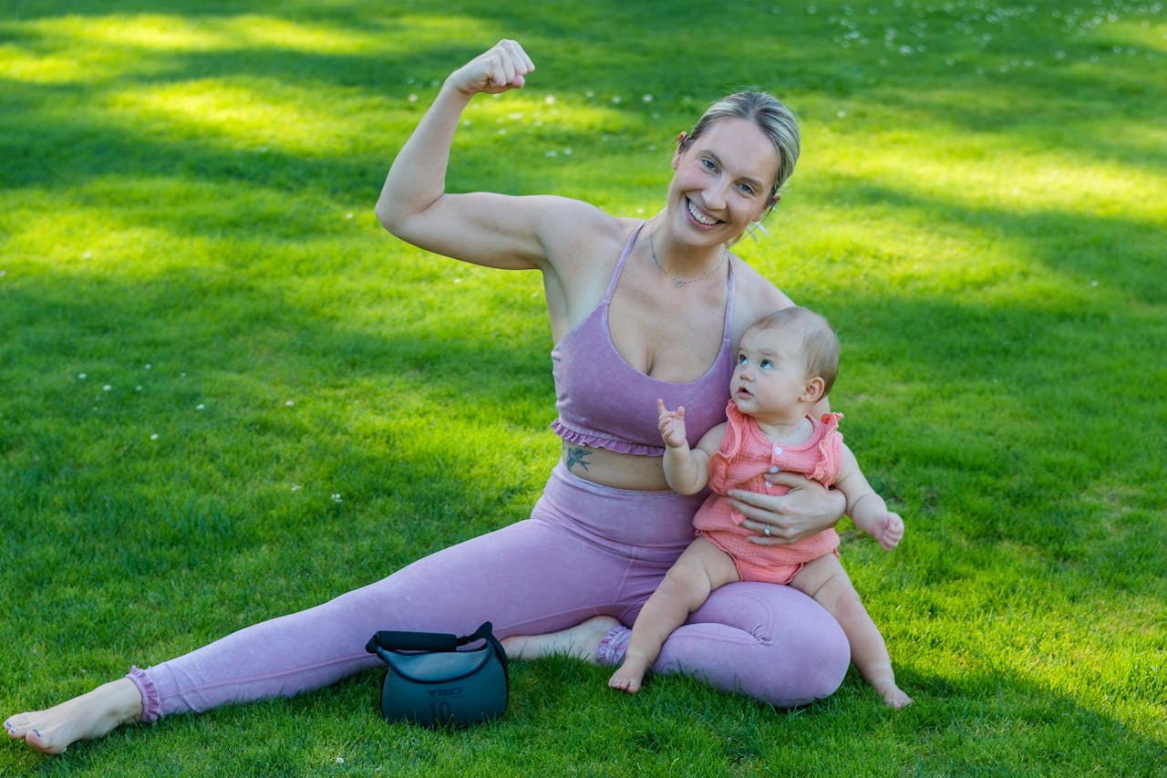 A smiling mother flexes playfully while sitting with her baby on a sunny lawn